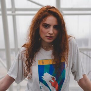 Portrait of a woman with long red hair wearing a white graphic t-shirt, looking at the camera in a bright indoor setting.